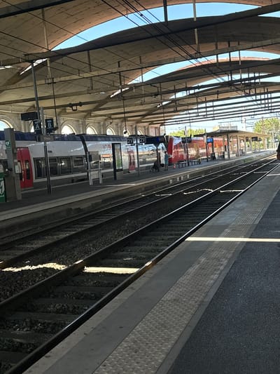 This is a vertical, eye-level medium shot of a train at a station in Nîmes, France. The train is primarily white with red accents on the front and rear carriages. It is stationary at the platform, suggesting passengers may be boarding or disembarking. A few people are visible on the platform, some appearing to be passengers and others possibly station staff. One person in the foreground is wearing a light blue shirt and shorts.

The scene is under a large, arched canopy supported by concrete pillars, typical of a European train station. The sky above is clear and blue, indicating fair weather and likely daytime. Sunlight casts strong shadows through the canopy structure and reflects off the train's windows. The train tracks run along the right side of the image, with ballast visible between the rails. The platform edge is marked with a textured, grey surface, designed for safety.

Signs are visible on the platform. One sign clearly displays "Nîmes" with an arrow, confirming the location. Other signs show letters and numbers, such as "A", "B", and "2V", likely indicating platform or track designations. There is also a sign that appears to be for "DAE" with an arrow. The overall atmosphere is one of a busy, functional transport hub on a bright day.