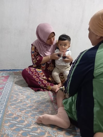 An indoor scene in Bulukandang, Indonesia, features three individuals on a multi-colored patterned rug against a plain light-colored wall. An adult female, wearing a light purple hijab and a maroon dress patterned with gold, brown, and light blue motifs, sits cross-legged, supporting an infant. The infant, wearing a light-colored top and pants, holds two small dark blue cylindrical objects in its raised hands and gazes towards the right. An adult, partially visible from the right side, wears an orange head covering and a dark blue long-sleeved garment with a prominent light green vertical stripe, with a light pink sock covering their right foot. The female on the left is positioned to interact with the infant, while the infant appears to be observing the third individual on the right.