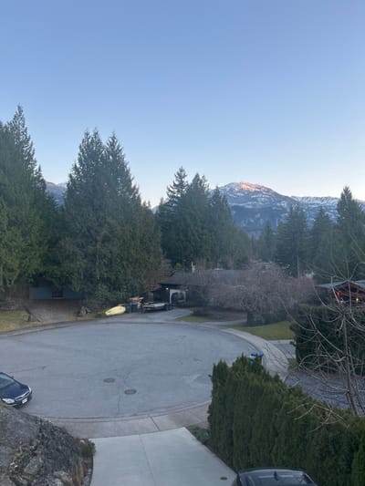 A residential street curves gently, lined with evergreen trees and homes. A construction vehicle and various equipment are parked along the side. In the distance, a mountain range is visible under a clear sky. A dark-colored car is parked near a rocky outcrop in the foreground. The scene appears to be captured from an elevated perspective, offering a view down the street.