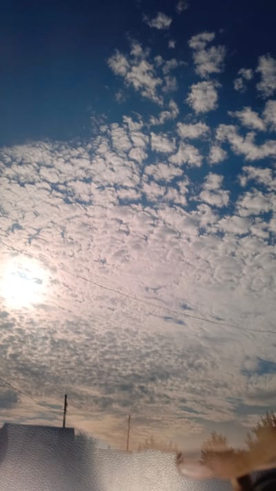 A vertical, eye-level shot captures a sky filled with numerous small, white clouds. A bright sun is visible in the upper left quadrant, partially obscured by some of the clouds. A thin, dark horizontal line, likely a power line, stretches across the mid-section of the frame. In the lower portion of the image, the top of a building or structure with a textured surface is visible, along with two utility poles. Trees are faintly discernible in the background. The scene appears to be an outdoor environment, possibly in a suburban or rural area, given the presence of utility poles and the building. The lighting suggests it is either early morning or late afternoon. The location is identified as the city of Zuy, Russia.
