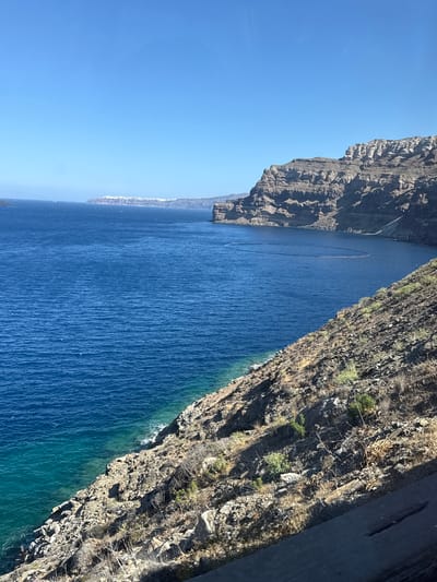 This media file captures a scenic view of the Aegean Sea and the dramatic cliffs of Santorini, Greece. The image is oriented vertically, presenting a wide expanse of deep blue water that transitions to a clearer, turquoise hue near the shore. The water exhibits a textured, wavy surface, indicative of gentle movement.

Dominating the right side of the frame is a steep, rocky cliff face, covered in sparse, dry vegetation and scree. The cliff's rugged texture and sharp inclines suggest a volcanic origin, characteristic of Santorini's caldera. A paved road is visible running along the top of the cliff on the far right.

On the left side, a distant coastline is visible, characterized by a dense cluster of white buildings, likely the iconic villages of Santorini, perched precariously on the cliff edge. Further out to sea, several small, dark objects are scattered, possibly boats or buoys, and a distinct, elongated dark line stretches across the water, potentially a floating barrier or a boat wake.

The sky is a clear, vibrant blue, suggesting a bright, sunny day. The lighting indicates that the sun is high in the sky, casting shadows that highlight the contours of the cliffs. The overall impression is one of natural grandeur and the stark beauty of the volcanic island landscape meeting the vast sea. No people are directly visible, and no text is present in the image.
