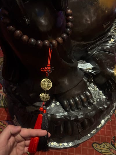 This media file features a close-up, overhead view of a dark, possibly bronze, statue, likely of a religious or cultural figure, given the presence of prayer beads draped around it and coins scattered around its base. The statue appears to be large and rests on a patterned red surface, possibly a rug or carpet, with a hint of a yellow and black butterfly design visible on the right side. The statue itself is detailed, with clearly visible fingers and toes, suggesting it might be a depiction of a deity or a revered person.
Draped across the statue's arm is a string of dark, polished wooden beads, characteristic of rosaries or prayer malas. Attached to the beads is a decorative tassel with red and black threads, and a small, round, golden coin or medallion. This coin has an intricate design featuring what appears to be a human figure and possibly some script. Another smaller, ornate golden charm is also attached to the tassel.
Scattered around the statue's feet and hands are numerous small, silver-colored coins, suggesting a practice of offering donations or wishes. A small white slip of paper, with some visible text that appears to be a voucher, is tucked near the statue's hands. A human hand is visible in the lower right corner of the frame, with a finger extended towards the statue's tassels, implying interaction with the object. The lighting suggests an indoor setting, possibly a temple, shrine, or a place of worship. The overall scene evokes a sense of reverence, tradition, and perhaps personal devotion. The location context provided is Shkotovo, Russia, which might imply the cultural or religious context of the statue within that region.