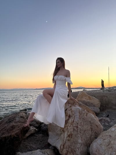 A young woman with long, auburn hair, wearing an off-the-shoulder white dress, sits on large rocks by the sea at sunset. The dress has a fitted bodice and a flowing, floor-length skirt that is split to reveal her legs. Her left leg is bent, with her foot resting on a rock. The woman is looking to her right, her gaze directed towards the horizon.
The background features a calm sea with gentle waves, meeting a shoreline with more rocks and some distant landmasses. The sky is a gradient of soft blues and oranges, indicating sunset. A sliver of a moon is visible in the upper left quadrant of the sky. In the middle ground, on the rocky shoreline, two figures are visible, one appearing to be fishing. A tall, slender pole, possibly a mast or a light pole, stands on the right side of the image. The scene suggests a peaceful, picturesque setting. The location is the coast of Paleo Faliro, Greece.