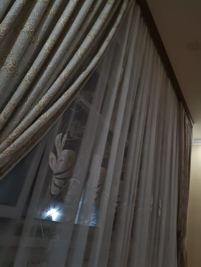 The image depicts a window covered by two layers of curtains. The inner layer is a sheer white curtain with a subtle floral pattern, while the outer layer is a heavier, textured beige curtain with a more prominent embossed pattern. The curtains are drawn, partially obscuring the view outside. A bright light source is visible through the sheer curtain, suggesting it may be nighttime or the room is dimly lit. The overall impression is one of domestic interior decor. The scene is located in the city of Ucar, Azerbaijan.