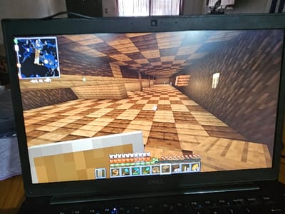 A laptop displays a Minecraft game. The in-game perspective shows a long corridor with alternating light and dark wood plank flooring and dark wood plank walls. A map is visible in the top left corner. The player's inventory is displayed at the bottom of the screen showing resources and items. The game is being played on a Dell laptop, with a black chassis and visible keyboard.