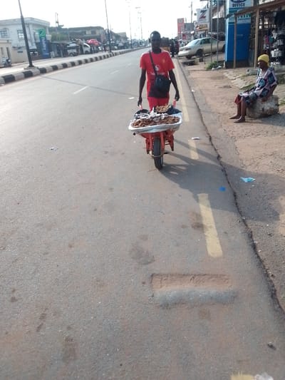 The image depicts a street scene in Orlu, Nigeria. A man in a red t-shirt and shorts is pulling a red wheelbarrow filled with food items. He is walking on the paved road, near the edge. An older woman wearing a yellow headscarf and patterned clothing is sitting on a rock on the side of the road. In the background, there are buildings, shops, and parked vehicles. A bank (Fidelity) logo is visible in the background. The road has a faded yellow line marking. The overall light suggests it is daytime.