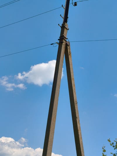 This media file shows a concrete power pole against a bright blue sky with scattered white clouds. The pole is V-shaped, with two concrete legs angled upwards and joined at the top. Wires are strung between the poles, supported by insulators attached to the top of the pole. The lighting suggests it is daytime, likely midday given the bright sunlight and clear sky. The presence of a power pole indicates an outdoor, possibly rural or suburban, setting. The overall mood is serene and calm, dominated by the expansive sky and the functional structure of the power pole. There is no visible text in the image.