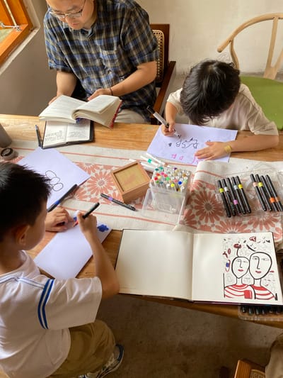 An overhead shot shows a man in a plaid shirt reading a book at a wooden table. He is sitting near a window with a view of greenery outside. Two children are drawing at the same table. One child, in a light-colored shirt, is drawing on a white sheet of paper with markers. The other child, in a white and blue striped shirt, is also drawing on paper, with a red marker in hand. Various art supplies, including markers and a sketchbook filled with abstract drawings of faces, are spread across the table. The scene takes place indoors, likely in a home or a cafe, suggesting a relaxed and creative atmosphere. The image does not provide enough information to determine the exact location in Dujiangyan City, China.