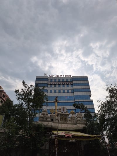 This image presents a detailed view of an urban landscape in Hyderabad, India, characterized by the juxtaposition of modern architecture and traditional religious structures under an overcast sky.

Dominating the upper portion of the frame is the multi-storied "MY HOME HUB" building, a contemporary high-rise featuring a facade composed of blue and white reflective panels or windows arranged in horizontal bands. The building's name, "MY HOME HUB," is prominently displayed in red, illuminated letters on its rooftop, with what appear to be communication antennas or structures nearby. Below this signage, on a lower floor, additional faint text or a logo, possibly "ICTOP" or "INSTITUTE OF TECH," is discernible.

In the midground and foreground, a traditional Hindu temple structure made of light-colored stone is visible, partially obscured by dense green foliage from several mature trees. The temple exhibits intricate carvings of deities and traditional motifs, with distinct spires and a golden-colored, wheel-like religious symbol adorning one of its upper sections. A yellow and red tarp or canopy provides shade over the lower, more obscured part of the temple, suggesting a temporary setup or a small vendor stall. A slender pole stands upright in front of the temple.

On the far left, an older, brownish-red building with multiple windows can be seen partially behind the trees. The sky is largely overcast, filled with various shades of grey and white clouds, suggesting a cloudy day with diffused lighting conditions. No individuals are clearly visible in the scene. The overall environment reflects a bustling urban area where modern commercial development coexists with established cultural and religious landmarks, specifically indicative of areas like Madhapur or Hitech City in Hyderabad, given the identifiable "MY HOME HUB" building.