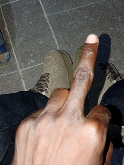 The image shows a close-up view of a person's right hand, with their index finger extended. The hand is positioned in the foreground, obscuring much of the rest of the scene. The person's skin tone is dark. The extended index finger appears to be the focal point, with visible skin texture and a light-colored fingernail.
The person is wearing dark pants and a light-colored shoe, likely a sneaker, which is visible in the background along with another similar shoe. The shoes have laces and are a neutral color, possibly tan or beige. The ground appears to be tiled, with visible grout lines. The lighting suggests it could be daytime, with a shadow cast by the extended finger onto the ground.
The image captures a moment of posed gesture, with the extended index finger possibly signifying a point, an insult, or some other form of communication. There is no visible text in the image. The setting appears to be an outdoor or semi-outdoor space with a tiled floor. The overall impression is casual, with the focus on the hand gesture and the immediate surroundings. Given the provided location context of Ukpor, Nigeria, the scene likely takes place there.