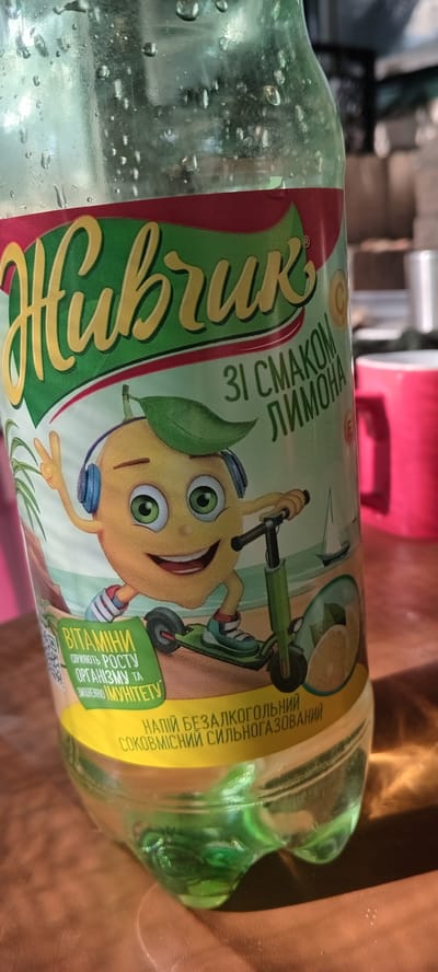 The image is a close-up, eye-level shot of a bottle of "Живчик" (Zhyvchyk) soft drink, a Ukrainian brand. The bottle has a green and yellow label featuring a cartoon lemon character wearing headphones and riding a scooter. Text on the label is in Ukrainian and reads "ЗІ СМАКОМ ЛИМОНА" (with lemon flavor), "ВІТАМІНИ СПРИЯЮТЬ РОСТУ ОРГАНІЗМУ ТА ЗМІЦНЕННЮ ІМУНІТЕТУ" (vitamins promote the growth of the organism and strengthen immunity), and "НАПІЙ БЕЗАЛКОГОЛЬНИЙ СОКОВМІСНИЙ СИЛЬНОГАЗОВАНИЙ" (non-alcoholic juice-containing highly carbonated drink). The bottle is placed on a wooden surface, likely a table, outdoors, with blurred background elements that suggest a sunny day. In the background, part of a red mug and some vegetation are visible. The presence of condensation on the bottle indicates it is a cold beverage. The image does not contain any information about the city of Koviagi, Ukraine, or any specific locations within it.