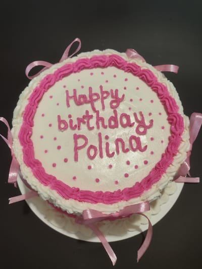 A white cake with pink polka dots and pink and white frosting borders is decorated with the words "Happy birthday Polina" in pink icing. Pink ribbons are tied around the cake. The cake sits on a white plate against a black background. The image was taken in Myadzyel, Belarus.