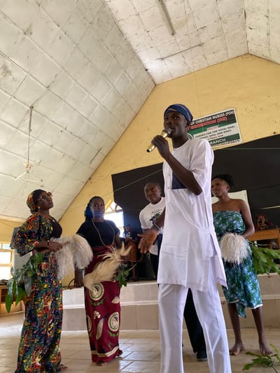 This image captures a gathering, likely a church event, in Dutsen Kato, Nigeria. Several people are present, including a young man speaking into a microphone, dressed in a white traditional outfit and a blue headwrap. He appears to be the focus of attention. Behind him, several women are also visible, some wearing colorful traditional attire. One woman in the background wears a blue and green patterned fabric, while another is dressed in a dark top and a vibrant, patterned skirt. The setting appears to be indoors, with white tiled walls and a yellow ceiling. A banner in the background reads, "OF CHRISTIAN NURSES (FCN) T GENERATION (NG)" with a motto and scriptural reference. The overall atmosphere seems formal yet celebratory, with the presence of traditional garments and what looks like a ceremonial element with white feathery objects held by some of the women. The lighting suggests it could be daytime, with natural light possibly coming from an unseen window. The image is oriented vertically.