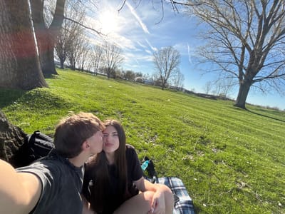 The image shows a sunny outdoor scene. Two people are close to each other, possibly kissing or embracing. They are sitting on a plaid blanket in a grassy area. One person has long, dark hair and is wearing a black shirt. The other person has shorter, light brown hair and is wearing a grey shirt. There is a backpack next to the person in the grey shirt. A water bottle and other items are near the blanket. The background shows a bright blue sky with sparse, bare trees lining the horizon. It is not possible to verify the location as Beli Brod, Bulgaria from this image alone.