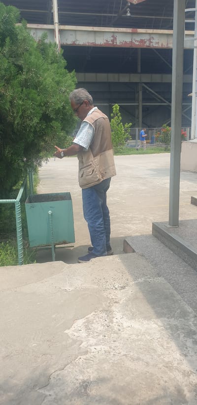 A man with gray hair and glasses stands on outdoor steps, holding a cigarette and looking down at it. He is wearing a light-colored patterned shirt, a tan vest with multiple pockets, and blue jeans. Beside him, to his left, is a light green metal trash can, partially obscured by a large evergreen tree. The ground is paved with concrete, and in the background, a large industrial building with a metal roof structure is visible. Further in the background, to the right, a chain-link fence runs along the edge of the paved area, and beyond the fence, two other individuals can be seen walking, one in a blue shirt and beige pants. The scene appears to be an outdoor area within the city of Rooppur, Bangladesh.