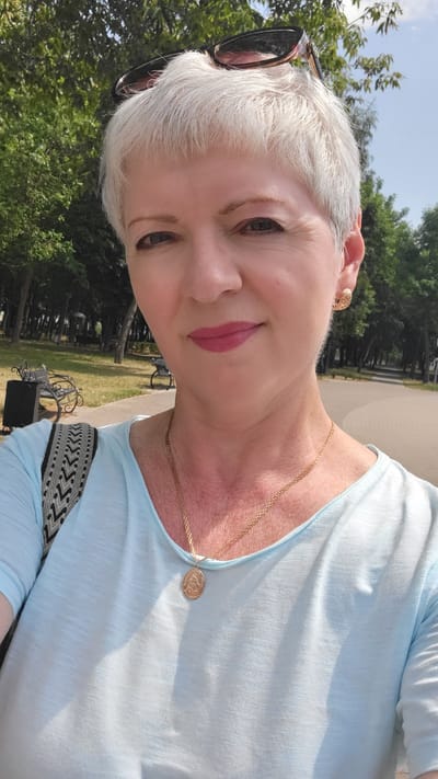 The media file is a portrait of a woman in an outdoor setting. She has short, white hair, is wearing a light blue top, a gold necklace with a pendant, and sunglasses pushed up on her head. Her makeup includes pink lipstick. The background shows greenery and trees, suggesting a park or natural area, with what appears to be a bench in the distance. The lighting indicates it is daytime, likely sunny. The overall impression is casual and relaxed. The provided location context is Belaazyorsk, Belarus.