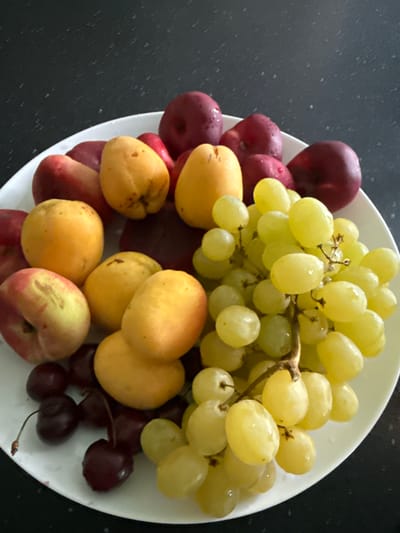 A white ceramic plate, filled with a colorful assortment of fresh fruits, is presented on a dark, speckled surface, possibly a countertop. The fruits include a large bunch of plump, pale green grapes, interspersed with several bright yellow apricots or pluots. Scattered among the larger fruits are deep red cherries, some still attached to delicate stems. The plate also contains several deep red plums or nectarines, their smooth skins glistening with water droplets, suggesting they have been recently washed. The overall impression is one of a vibrant, healthy fruit selection, possibly prepared for a meal or as a refreshing snack. The lighting appears to be natural, casting soft shadows and highlighting the dewdrops on the fruit, indicating it might be daytime. No people or text are visible in the image. The context of Malmyzh, Russia, does not provide specific visual cues within the image itself, but suggests a domestic setting.