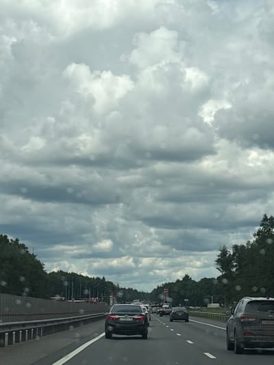 The image depicts a view from a vehicle traveling on a highway, likely in the city of Shcheglovo, Russia, given the context of the provided crops. The sky is filled with dense, gray, and white clouds, suggesting an overcast or potentially stormy day. The highway is multi-lane, with several cars visible in the frame. In the foreground, on the right side of the highway, a dark-colored SUV is prominent. Further ahead on the highway, other vehicles are in motion, and on the left side of the road, a concrete barrier separates the lanes from the adjacent landscape. Beyond the barrier, trees and what appear to be streetlights line the side of the road. Some of the trees are dense and tall, indicating a natural or forested area bordering the highway. There are also some distant structures that might be related to infrastructure or services along the route. The image has a slightly blurry quality, possibly due to motion or water droplets on the windshield from which the photo was taken. No people are directly visible in the frame.
