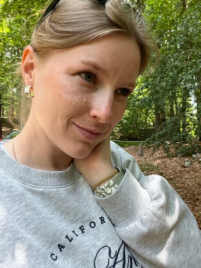 A young woman with blonde hair and green eyes is pictured outdoors, resting her chin on her hand. She is wearing a light gray sweatshirt with the word "CALIFORNIA" embroidered on it in dark blue lettering. She has on hoop earrings with a yellow and white design, and a thin gold chain necklace. Her cheek is adorned with small, scattered glitter accents. The background is a natural environment with trees and fallen leaves on the ground, suggesting a park or wooded area. The location is not in Mali Bostur, Montenegro.