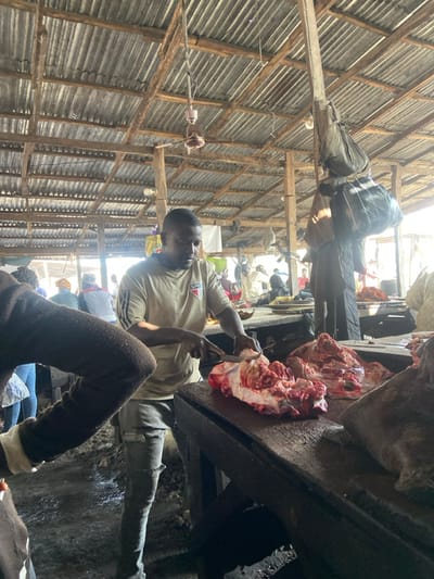 The image depicts an operational market scene within Kofar Mazugal, Nigeria. A male individual, identified as a butcher, stands center-right, actively cutting raw meat. He has a dark complexion and wears a light-colored t-shirt with "SPF" visible on the chest, along with dark, distressed jeans. He holds a large knife in his right hand, severing a substantial piece of red meat, which includes muscle, fat, and bone, on a dark wooden butcher block. An additional large cut of raw meat rests on the same surface to his right. The market structure features a corrugated metal roof supported by a visible framework of wooden beams. A light-colored ceiling fan hangs from the roof structure. A bare incandescent light bulb is suspended from a vertical wooden post, from which several dark, bundled objects are also hanging. The ground in the foreground appears dark and irregular. The upper arm and shoulder of a foreground figure, clothed in dark, possibly textured fabric, are visible on the left. Several other individuals are discernible in the mid-ground and background, participating in various market activities.