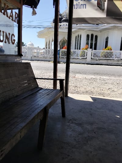 A weathered wooden bench sits on a concrete surface, partially shaded. In the background, a white building with intricate architectural details, including arched windows and a dome, is visible. A white fence lines the perimeter of the building. A paved road stretches in front of the building. Overhead power lines are suspended in the bright sky. The overall scene suggests a roadside view of a religious or public building.