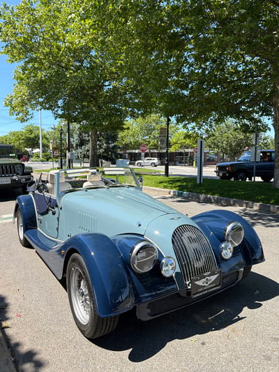 A two-toned light blue and dark blue vintage Morgan car is parked on an asphalt road in front of trees with green leaves and a clear blue sky. The car has a prominent chrome grille and two round headlights. A license plate on the front of the car reads "MORGAN FORT LAUDERDALE". Behind the Morgan, a dark green Jeep Wrangler is parked. In the background, there is a street with other vehicles and buildings. The scene appears to be in a town or city.
