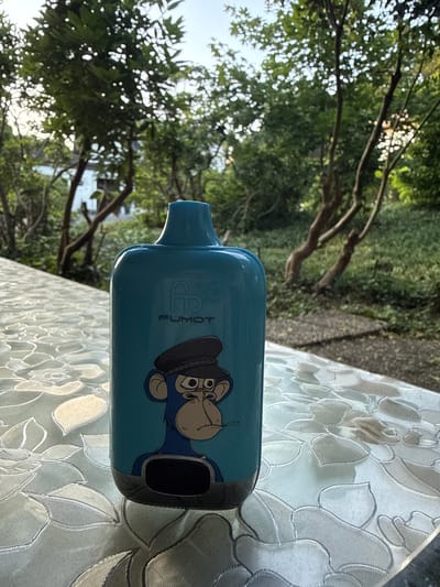 A light blue vape device with an ape graphic and the brand name "FUMOT" printed on it sits on a patterned tablecloth. The device is positioned slightly off-center to the right. In the background, trees and foliage are visible, suggesting an outdoor setting. The lighting indicates it is daytime, likely late afternoon or early evening due to the soft shadows. No specific location in Herford, Germany, can be identified from the image.