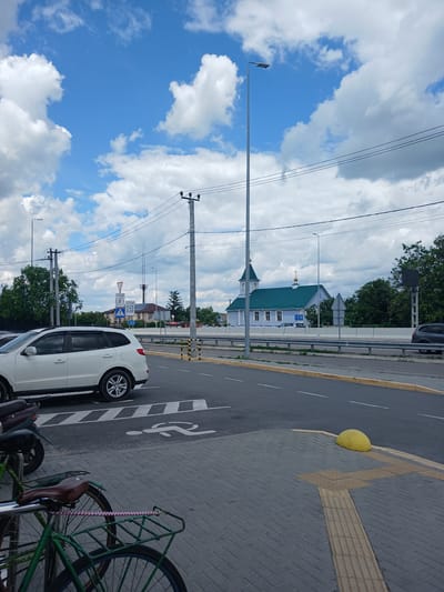 This image captures a street scene in Hrebinky, Ukraine, under a partially cloudy blue sky. In the foreground, a green bicycle with a brown seat and a basket is parked on a gray brick sidewalk. Adjacent to it, another bicycle and a dark-colored motorcycle are also parked.

A white SUV is positioned on the asphalt road, partially obscuring a crosswalk marked with white stripes. A symbol of a walking person is painted on the crosswalk. A yellow bollard is located on the sidewalk to the right of the crosswalk.

Further down the road, a light blue church with a green roof and gold-colored crosses stands prominently. Several traffic signs are visible, including a yield sign, a sign indicating a speed limit of 5, and a parking sign. Power lines crisscross the sky, supported by utility poles. In the background, trees and buildings line the horizon, with a concrete barrier separating the road from the area behind it. The overall scene depicts a typical urban environment with elements of transportation and community infrastructure.
