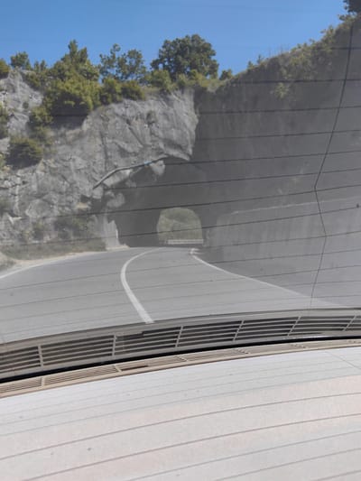 The image shows a view of a road through a tunnel, as seen from inside a vehicle. The road curves slightly to the left as it enters the tunnel. A single white line divides the road. The tunnel appears to be carved through a rocky hillside with vegetation on top. The image is partially obscured by what appears to be the rear window of the vehicle, including the defroster lines and the rear deck. The precise location in Bilka, Bulgaria cannot be determined.