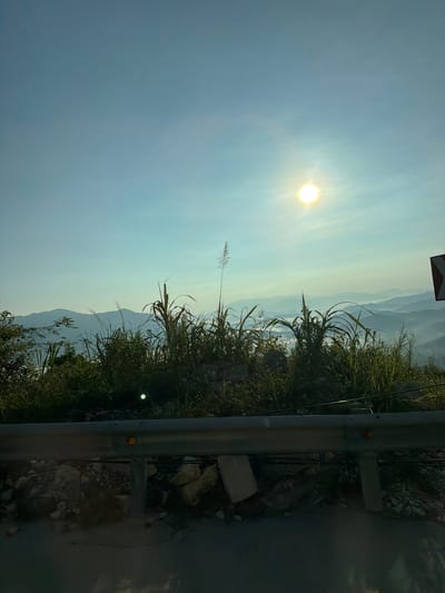 The image depicts a scenic view of mountains and a hazy valley, likely from an elevated vantage point. The sun is high in a clear blue sky, casting a bright glow. Tall grasses and foliage are in the foreground, partially obscuring the view of the landscape beyond. A metal guardrail runs horizontally across the lower portion of the image, suggesting the photograph was taken from a road or pathway. In the far distance, layers of mountains recede into the haze. The overall impression is of a tranquil, natural environment, possibly captured during sunrise or sunset given the soft light and atmospheric conditions. The location is identified as Nam Khanh Vinh, Vietnam.