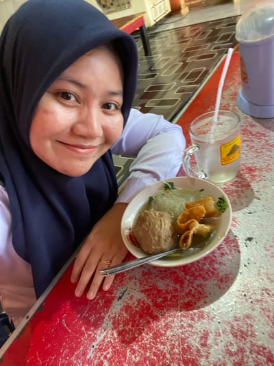 A person wearing a dark blue hijab and a white shirt is taking a selfie in an outdoor setting. They are smiling and looking at the camera. To their right, there is a bowl of soup with meatballs and noodles, and a glass of iced water with a straw. The table is red and appears to be made of metal. In the background, there is a patterned floor and a glimpse of a purple dispenser. The setting appears to be a casual dining area, possibly a food stall or a simple restaurant. The image was taken in the city of Sungai Sipai, Indonesia.
