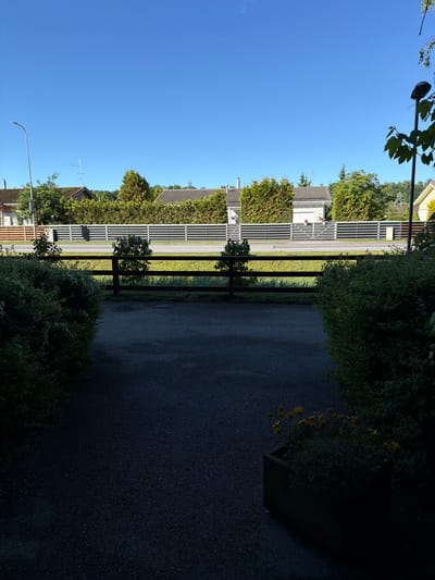 The image depicts a residential street scene in Tabasalu alevik, Estonia. In the foreground, a dark asphalt driveway is partially shaded, with a wooden planter box containing yellow flowers visible on the right. A wooden fence runs horizontally across the mid-ground, separating the driveway from a grassy area. Beyond the fence, a grey horizontal slat fence lines the edge of a road, with a sidewalk in front of it. Residential houses with pitched roofs and manicured evergreen hedges are visible behind the slat fence, set against a clear blue sky. A street lamp is present on the left side of the frame. There are no people or animals clearly visible in the image. The scene appears to be set during daylight hours with bright sunlight casting shadows.