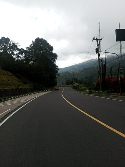 The scene depicts a winding asphalt road in Deothang, Bhutan, under an overcast sky. The road features a continuous yellow centerline and a solid white line marking the right shoulder, which abuts a patterned concrete curb. To the left, a terraced hillside secured by stone retaining walls is densely covered with green trees and vegetation. A light-colored compact van or car is observed in the mid-distance, traveling away from the viewer on the left lane of the road as it curves to the right. On the right side of the road, multiple utility poles support an array of overhead power and communication lines. Beyond this infrastructure, densely forested mountains rise, extending into the distance, partially obscured by clouds and mist, with faint signs of distant structures on their slopes. No individuals are visible.