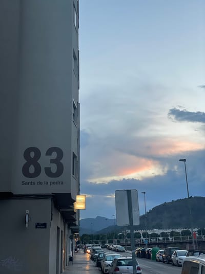 The image depicts a street scene in Sagunto, Spain, with a large building occupying the left side of the frame. The building has the number "83" prominently displayed in dark grey on its facade, with the text "Sants de la pedra" below it. A smaller sign for "PODOCENTRE" is also visible on the building. A street sign indicating "Carrer Benifairó" is attached to the building's corner. The street is lined with parked cars, and pedestrians are visible walking on the sidewalk. In the background, a hill covered in trees rises above a bridge or overpass, with a cloudy sky overhead. The lighting suggests it is either early morning or late evening, with some artificial lights illuminating the street.