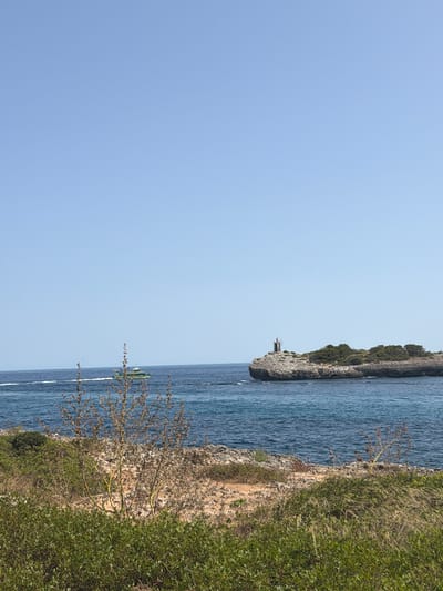 The image depicts a coastal landscape in Manacor, Spain. The scene is dominated by a clear, bright blue sky, which takes up the majority of the frame. Below the sky, the deep blue sea stretches out, with gentle waves indicating a calm day. In the distance, a small, rocky island juts out from the water, topped with sparse vegetation and a small lighthouse. To the left of the island, a green boat with white trim is moving across the water, leaving a visible wake. The foreground of the image is filled with rocky terrain and various types of greenery, including bushes and dry, wispy plants. The overall impression is one of a sunny day at a Mediterranean coast.