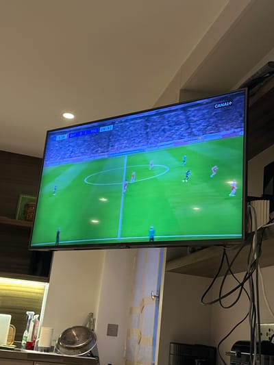 The image shows a television screen displaying a soccer game, specifically a match between Bayern Munich (BAY) and Paris Saint-Germain (PSG). The score is BAY 0-1 PSG, and the time is 3:58. The channel logo "CANAL+" is visible in the top left corner. The television is located in a kitchen area with cabinets, appliances, and a window. The location is suggested to be in Neuilly-sur-Seine, France, based on the French TV channel. There are no people clearly visible in the frame.