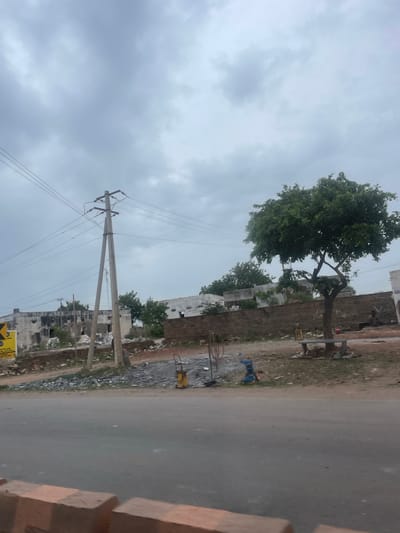 The image depicts a street scene in Bethamcherla, India, under a cloudy sky. In the foreground, a paved road runs horizontally across the frame. Beyond the road, there is a gravelly area with various objects, including construction materials like grey slate tiles, a yellow machine that appears to be a compactor, and a blue cart. A person is seated on a bench under a large, leafy green tree to the right of the frame. Behind the tree, there is a low, textured brown wall, and further back, several dilapidated buildings with broken windows and weathered facades are visible. A prominent concrete utility pole with multiple wires stretches vertically in the middle left of the image. The overall impression is one of an underdeveloped or developing area undergoing construction or renovation.