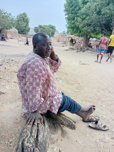 The image shows an adult man lying on a weathered log outdoors in what appears to be a rural area in Nata-Ala, Nigeria. He is wearing a patterned shirt, dark blue pants with a red stripe, and is barefoot. His sandals are placed next to his feet on the sandy ground. The man's head is turned slightly, and his hand is near his head as if he is resting. In the background, there are several people and structures that suggest a village setting. A group of children are walking down a path, and beyond them are simple buildings constructed of mud or similar materials. Trees are scattered throughout the scene. The ground is dry and sandy, consistent with a rural environment.