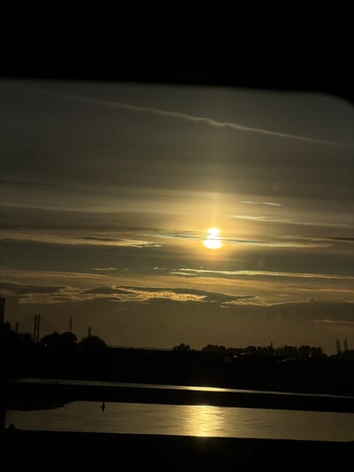 This image depicts a serene, dusky scene of a waterfront with a dramatic, hazy sunset. The sun, a brilliant golden orb, is partially obscured by clouds, creating a soft, diffused glow that illuminates the sky with streaks of yellow and white. The clouds themselves appear elongated and wispy, contributing to the ethereal atmosphere.

On the right side of the frame, a dark silhouette of a shoreline is visible, dotted with trees and a few man-made structures that appear to be piers or docks. The water in front of this shoreline reflects the golden light of the setting sun, creating a shimmering, textured surface. A lone bird is captured in silhouette, mid-flight over the water.

The left side of the image is dominated by a dark, almost black area, which may represent the foreground or a structure from which the photo was taken. There is a slight, soft glow emanating from the right edge of this dark area, suggesting it is not entirely opaque.

The overall mood is one of tranquility and quiet beauty. The time of day is clearly sunset, indicated by the low angle and golden color of the sun, and the soft, diffused light. The weather appears calm, with the hazy conditions contributing to the softened visual quality. No text is visible in the image. The location context suggests this is a waterfront area in Richmond, Canada.