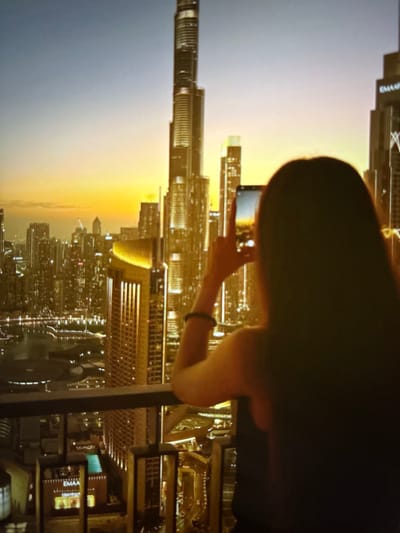 The image captures a woman from behind, standing on a balcony and taking a photograph of a cityscape with her smartphone. The cityscape is illuminated by the warm glow of a sunset, casting long shadows and highlighting the architectural details of numerous skyscrapers. The sky transitions from a deep blue at the top to a vibrant orange and yellow near the horizon. The woman, with her dark hair partially obscuring her face, is focused on her phone, her fingers positioned to operate the camera. She is wearing a dark top and a bracelet on her left wrist. The balcony railing is visible in the foreground, suggesting an elevated vantage point. The architecture in the background, with its sharp lines and glowing windows, is characteristic of a modern metropolis. The specific mention of Pori, Finland, is not supported by any visual cues in the image, as the skyline strongly resembles that of Dubai, known for its iconic skyscrapers like the Burj Khalifa.