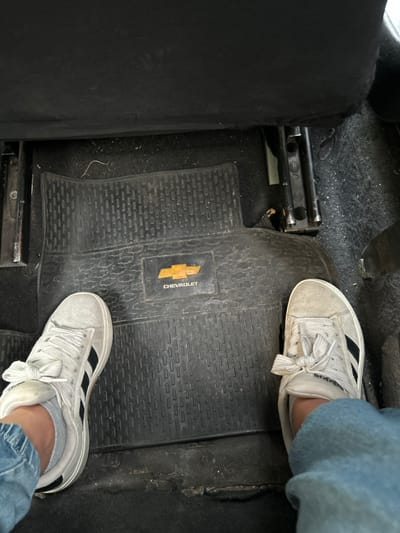 The scene depicts the rear passenger footwell of a Chevrolet vehicle in Ciudad del Libertador General San Martín, Argentina. A person's lower legs, clad in light blue denim jeans, and feet are visible. The individual wears light gray low-top sneakers with white laces and three black stripes on the lateral side of each shoe. Both feet rest flat on a dark, textured rubber floor mat, which features a gold Chevrolet bowtie emblem and the word "CHEVROLET" centrally. The bottom edge of a dark fabric car seat is visible at the top of the frame, with metal seat adjustment rails partially exposed on the left and right. The vehicle's floor is covered in dark carpet surrounding the mat. Minor dust and debris are present on the visible surfaces.