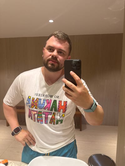 A male individual, appearing Caucasian with short dark brown hair and a trimmed beard, takes a mirror selfie in an indoor tiled room. He wears a white V-neck t-shirt featuring the mirrored, colorful text "NO WORRIES! HAKUNA MATATA!" and blue swim shorts. A black smartwatch is on his left wrist, and a light blue smartwatch is on his right. A thin gold chain is visible around his neck. He holds a dark smartphone with a triple-lens camera in his right hand, while his left hand rests on his hip. The room features light brown tiled walls, a lighter tiled floor, and a white ceiling with a single recessed light. A white sink basin and a metallic waste receptacle lid are partially visible in the foreground. A wooden bench is reflected in the background. The specific location within Zanzibar, Tanzania, is not discernible from the visual information provided.