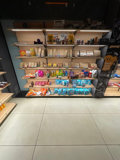 The image shows a well-lit grocery store shelf display in Sidhpur, India. The shelves are made of light-colored wood and are illuminated by strip lighting. The shelves are stocked with a variety of products, including packaged food items, toiletries, and cleaning supplies. Some of the brands visible include Tide, Ariel, and Pampers. There is a chalkboard-style illustration on the side of the display featuring food items and the text "Word Fails Food Speaks." The flooring is a light-colored tiled surface. The scene appears to be captured from a slightly elevated, wide-angle perspective, looking down and across the shelves. No people are present in the image.