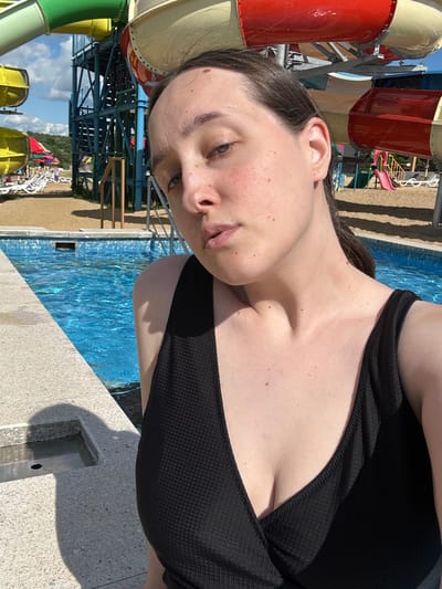 A young woman is posing for a selfie at a water park in Yubileyny, Russia. She is wearing a black swimsuit and is standing next to a pool. In the background, there are several water slides and lounge chairs. The sky is blue and there are some clouds. The woman has light skin and dark hair. She is looking at the camera with a neutral expression.