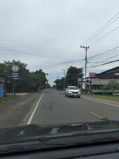 A cloudy, overcast day is depicted in this street-level, eye-level, point-of-view shot taken from a vehicle. The image captures a two-lane asphalt road that extends into the distance, flanked by a grassy shoulder on the left and more roadside vegetation on the right. A white SUV is in the right lane, traveling away from the camera. Numerous power lines and utility poles are strung across the sky, some crossing the road.
On the left side of the road, a prominent circular sign with a logo and text reads "AKOM JAYA BAKSO & RESTO". Below this, more text is visible, including "JAGONIAH MIE ACEH" and "0822 2828 8010". Adjacent to the sign is a building with a dark roof and some blue tarp material visible. Further down the road, on the right, a building with a thatched roof structure is present, with green pillars and a sign that reads "BAKSO CIPTON".
The overall scene suggests a semi-urban or rural setting in Meunasahlhok, Indonesia, along a well-traveled road with various businesses and infrastructure. The reflection on the vehicle's dashboard shows the environment outside, including trees and buildings.
