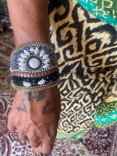 A high-angle close-up depicts a person's left forearm and hand, adorned with a decorative, wide bracelet, set against backgrounds of patterned fabric and carpeting. The bracelet features a black beaded base, white cabochon stones arranged in a circular pattern around a central larger stone, and gold chain edging. The person's skin exhibits visible veins and two small, stylized tattoos. The background includes a swatch of fabric with a black and beige animal print and another section of patterned fabric with green and blue tones. A portion of a multi-colored oriental rug is visible in the lower right corner of the frame. No specific information indicates this is Baan Nai Suan, Thailand.