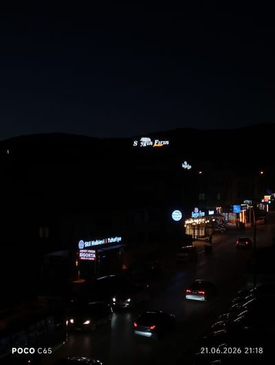 The image depicts a nighttime street scene in Körfez, Turkey, possibly a commercial area. Several illuminated signs are visible, including "Saygın Fırın" (Saygın Bakery) and "Stil Hobievi & Tuhafiye" (Style Hobby House & Haberdashery). A sign for "AHMET PEKER SİGORTA" (Ahmet Peker Insurance) is also present. Numerous cars are on the road, some with their headlights on, and others are parked along the street. The overall impression is of a relatively active street at night, with businesses open and traffic present. The dark sky suggests it is late at night or early evening. The timestamp in the lower right corner indicates the photo was taken on June 21, 2026, at 21:18.