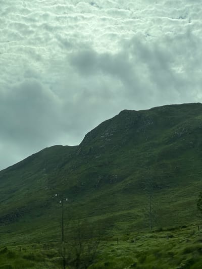 The image depicts a verdant hillside under a cloudy sky. The hillside is covered in lush green grass, with patches of rocky outcrops visible. In the mid-ground, a wooden pole with three lights on top stands tall, and further to the right, a large metal pylon is present. The sky is filled with textured, greyish-white clouds, with some darker clouds present towards the top. The overall impression is of a rural landscape in Ireland, possibly in the town of Ardnawark, characterized by its natural beauty and the presence of man-made structures that convey electricity across the land. There are no people visible in the scene.