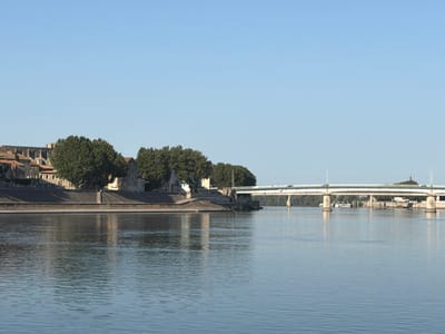 The image captures a wide view of the Rhone River in Arles, France, on a clear, sunny day. The river occupies the foreground, its surface rippled by a gentle breeze, reflecting the light blue sky. Along the left bank, an embankment lined with stone walls separates the river from the city. Above the embankment, a cluster of buildings with terracotta roofs and stone facades is visible, interspersed with large, mature trees. In the middle ground, a modern bridge spans the Rhone, supported by concrete pillars. Cars are visible on the bridge, indicating traffic. On the far bank of the river, more trees and buildings are present, with a distinctive red-domed structure visible in the distance. The overall impression is one of a tranquil, picturesque urban landscape.