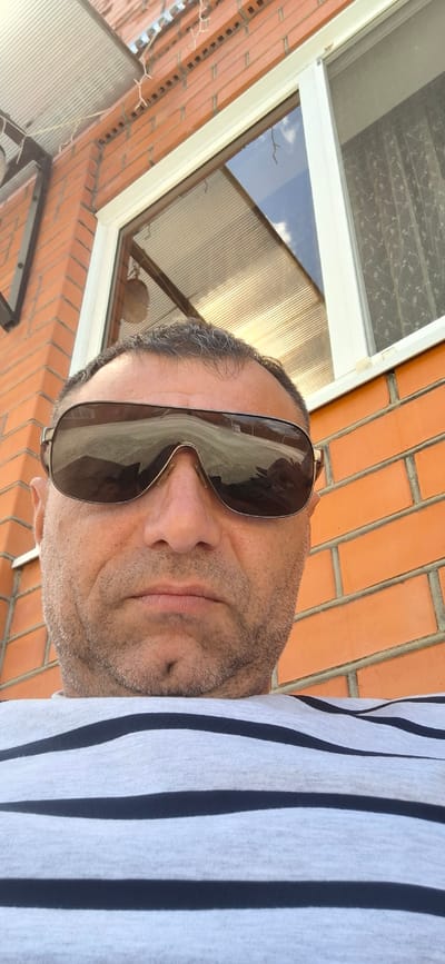 A man wearing sunglasses and a striped shirt is pictured from the chest up. He is outdoors, with a brick wall and a window visible behind him. The window reflects the sky and a covered walkway. The scene is likely in Kabekhabl, Russia.